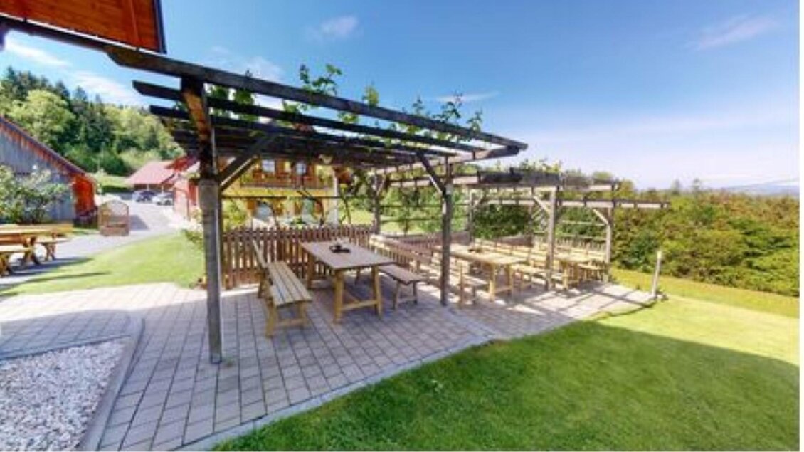 Eine gemütliche Terrasse mit Holzbank und Tisch unter einem Pergola-Baldachin. Im Hintergrund sind grüne Wiesen und ein klarer Himmel zu sehen. | © Helga & Robert | Panoramaschenke Kure
