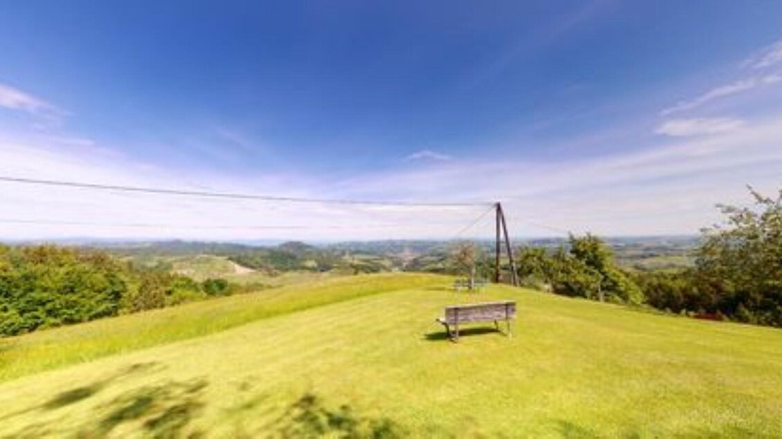 Ein weitläufiger grüner Hügel mit einer Bank und einem klaren blauen Himmel. Die Aussicht reicht über sanfte Hügel und Wälder. | © Helga & Robert | Panoramaschenke Kure