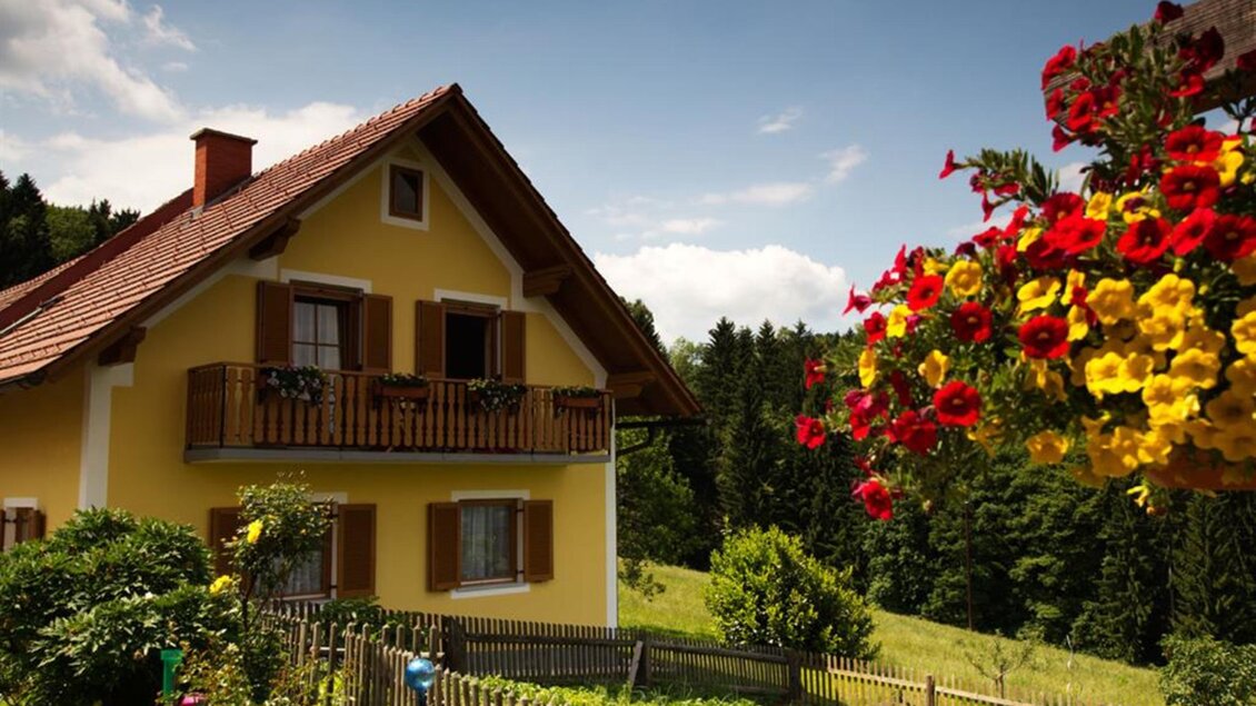 Ein gelbes Haus mit einem Balkon und schönen Blumen im Vordergrund. Die Landschaft ist grün und es gibt einen klaren blauen Himmel. | © Helga & Robert | Panoramaschenke Kure
