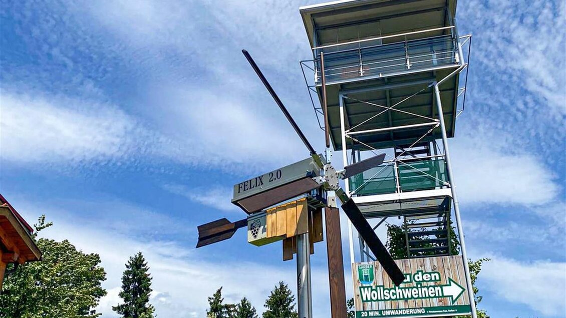 Ein Aussichtsturm steht im Vordergrund, umgeben von Bäumen und einer schönen Landschaft. Der Himmel ist klar und zeigt einige Wolken. | © Wein- und Genusswelt Garber
