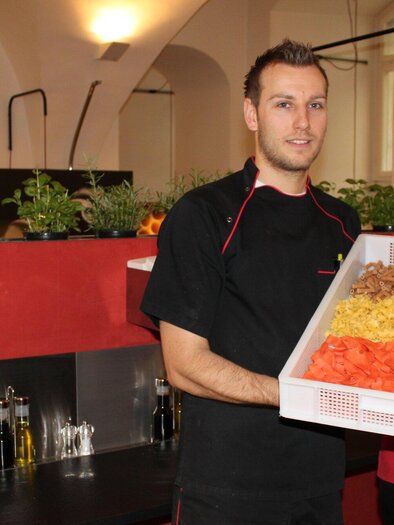 A man and a woman are standing in a restaurant holding a large box of colorful pasta. In the background, herb pots and kitchen utensils are visible. | © CMS