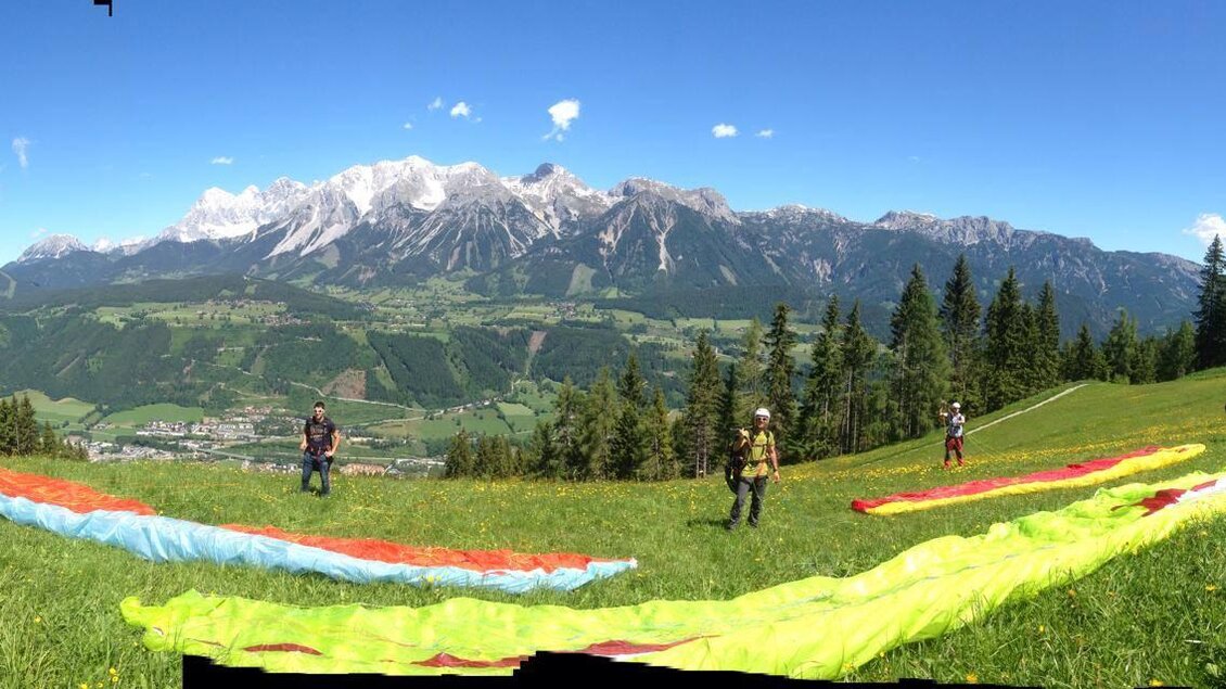 Ein schöner Bergblick mit grünen Wiesen und bunten Gleitschirmen. Die Landschaft ist klar und sonnig, mit schneebedeckten Gipfeln im Hintergrund. | © Flugschule Aufwind 