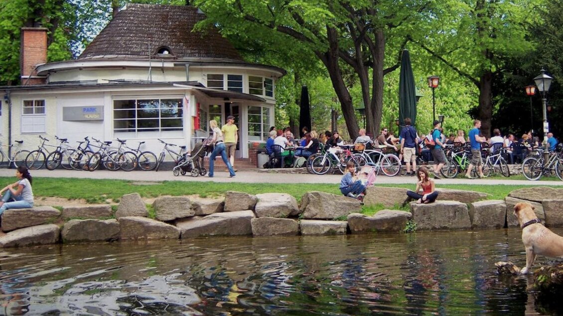 Ein lebhafter Park mit vielen Menschen, die an einem Café sitzen und Fahrräder parken. Im Vordergrund sieht man einen Hund am Wasser und Bäume im Hintergrund. | © Parkhouse - Huber
