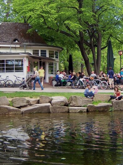 A lively park with many people sitting at a café and parking bicycles. In the foreground, you can see a dog by the water and trees in the background. | © Parkhouse - Huber