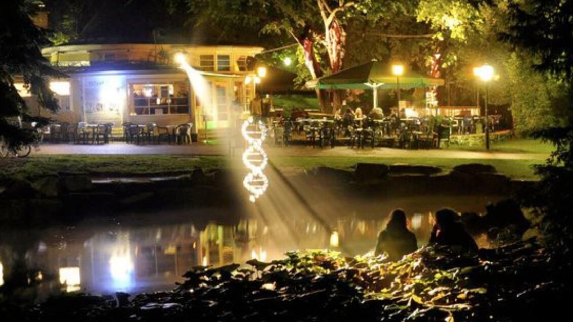 Ein malerischer Park bei Nacht mit beleuchtetem Restaurant und einer ruhigen Wasserfläche. Im Hintergrund sitzen zwei Personen am Ufer und genießen die Atmosphäre. | © Parkhouse - Huber
