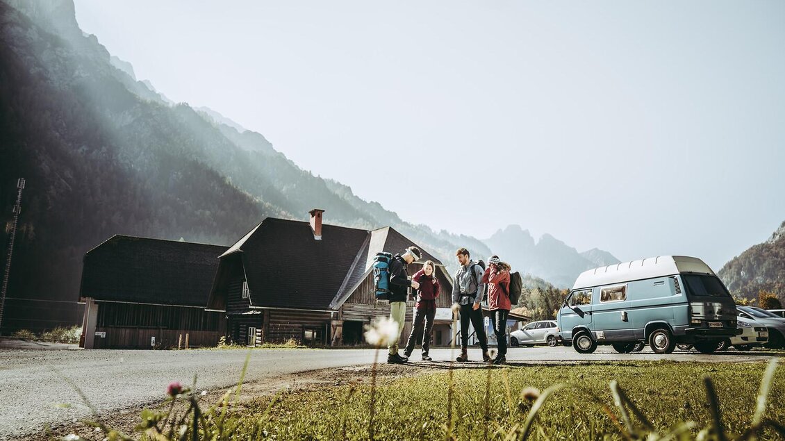 Eine Gruppe von vier Personen steht auf einer Wiese nahe eines Hauses und eines Wohnmobils. Im Hintergrund sind Berge und ein klarer Himmel zu sehen.
