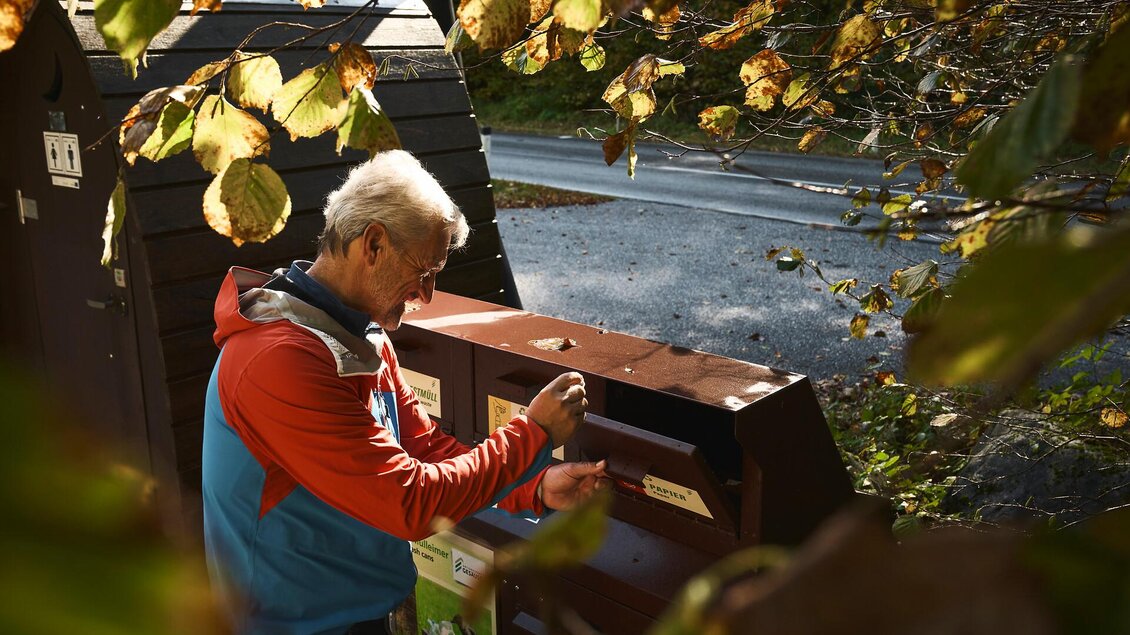 Ein Mann füttert einen Getränkespender in der Natur. Im Hintergrund sind herbstliche Blätter und eine ruhige Straße zu sehen.