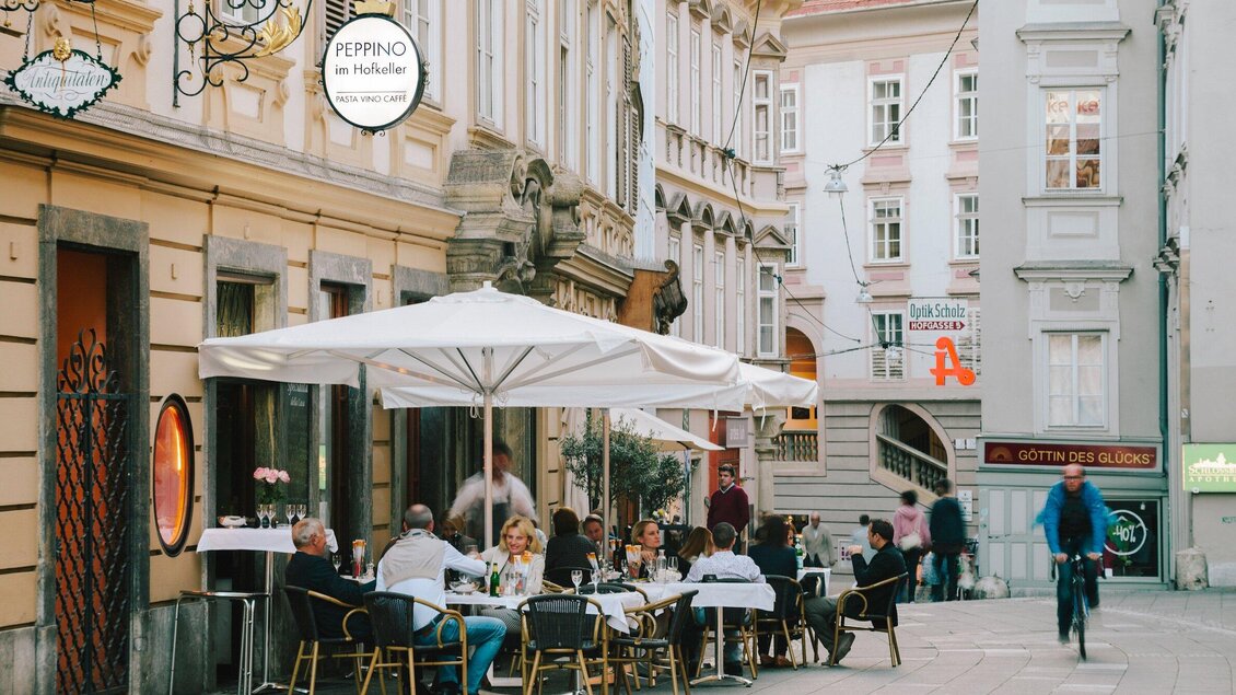 Eine belebte Straße mit einem Restaurant im Freien. Gäste sitzen unter einem Sonnenschirm und genießen ihr Essen. | © Peppino - Sebastian Reiser
