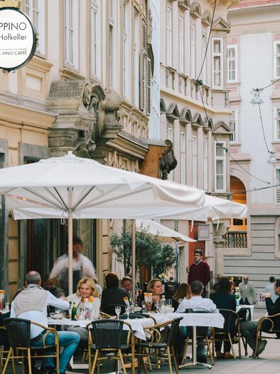 A busy street with an outdoor restaurant. Guests are sitting under a sunshade and enjoying their meal. | © Peppino - Sebastian Reiser