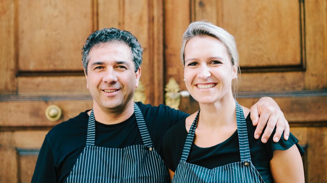 Ein lächelndes Paar steht vor einer hölzernen Tür. Beide tragen schwarze T-Shirts und gestreifte Schürzen. | © Peppino - Sebastian Reiser