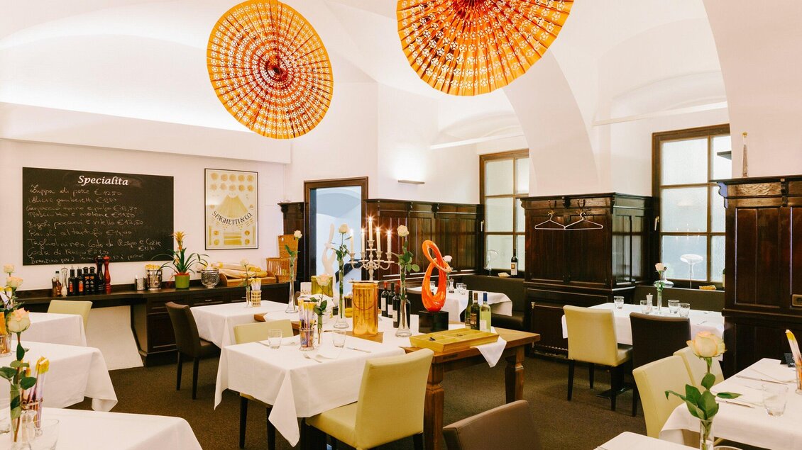 Ein elegantes Restaurant mit weißen Tischdecken und frischen Blumen. Die Wände sind in warmen Tönen gestaltet, und es hängen farbenfrohe Lampen von der Decke. | © Peppino - Sebastian Reiser