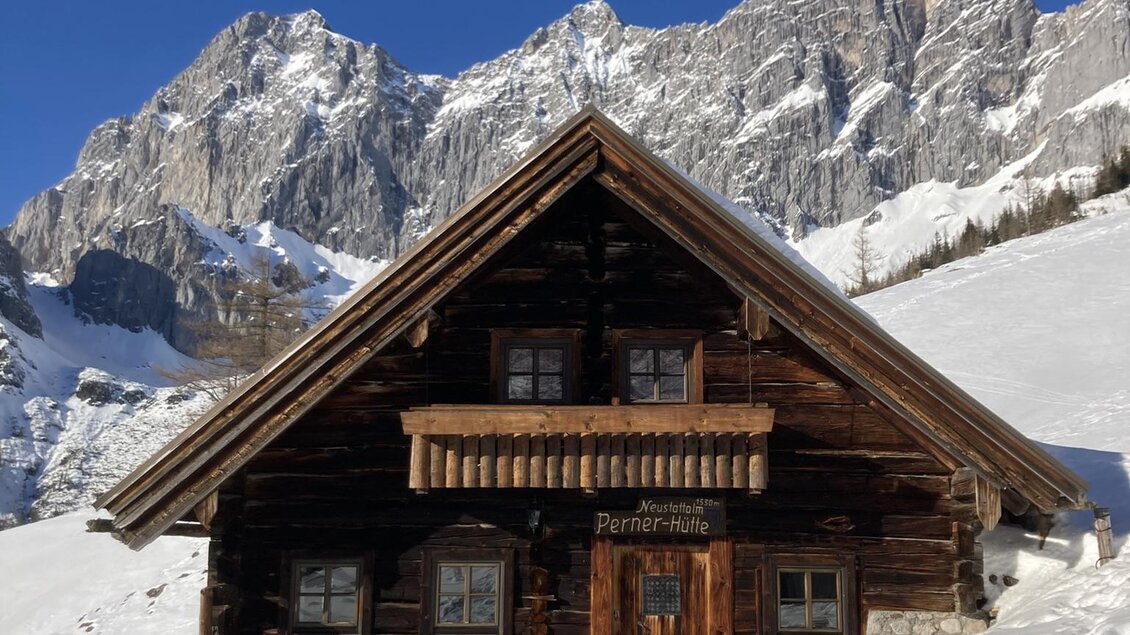 Eine gemütliche Holzhütte steht im Schnee, umgeben von majestätischen Bergen. Der Himmel ist klar und blau. | © Theresa Perner 