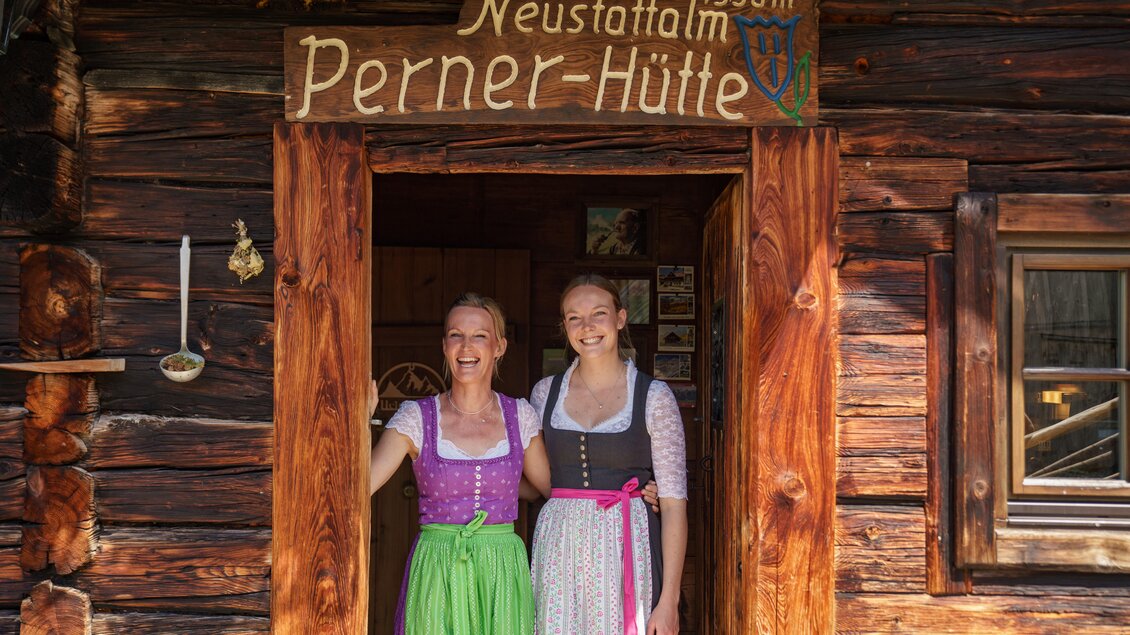 Zwei Frauen in traditionellen Trachten stehen vor der Perner-Hütte in den Bergen. Die Hütte ist aus Holz und hat ein Schild mit der Aufschrift „Neustattalm Perner-Hütte“ und einer Höhe von 1530 m. | © TVB Schladming-Dachstein/Christine Höflehner