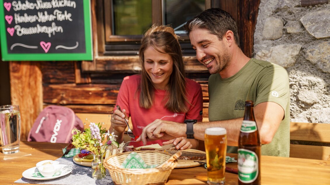 Ein Paar sitzt an einem Holztisch und genießt ein gemeinsames Essen im Freien. Auf dem Tisch befinden sich leckeres Essen, zwei Biergläser und eine schöne Blumendekoration. | © TVB Schladming-Dachstein/Christine Höflehner