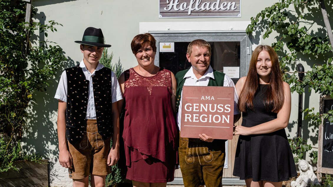 Eine Gruppe von vier Personen steht vor einem Laden mit dem Schild „Hafladen“. Sie tragen traditionelle Kleidung und halten ein Schild mit der Aufschrift „Genuss Region“. | © Martina Siebenhandl