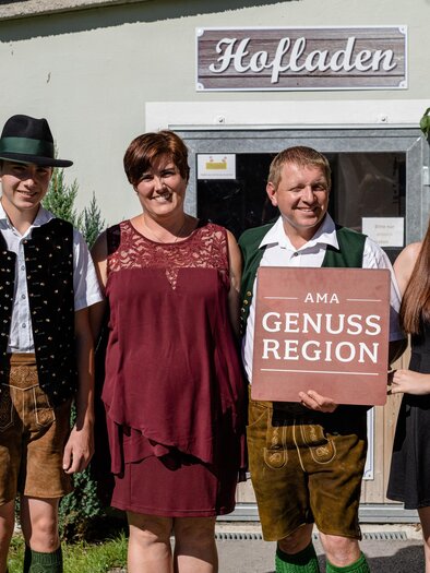 A group of four people stands in front of a store with the sign "Hafladen". They are wearing traditional clothing and holding a sign that says "Genuss Region". | © Martina Siebenhandl