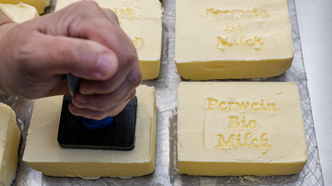 Eine Hand stempt eine Marke auf Butterstücke. Die Butter ist in gleichmäßigen Blöcken angeordnet und trägt Aufschriften. | © Martina Siebenhandl