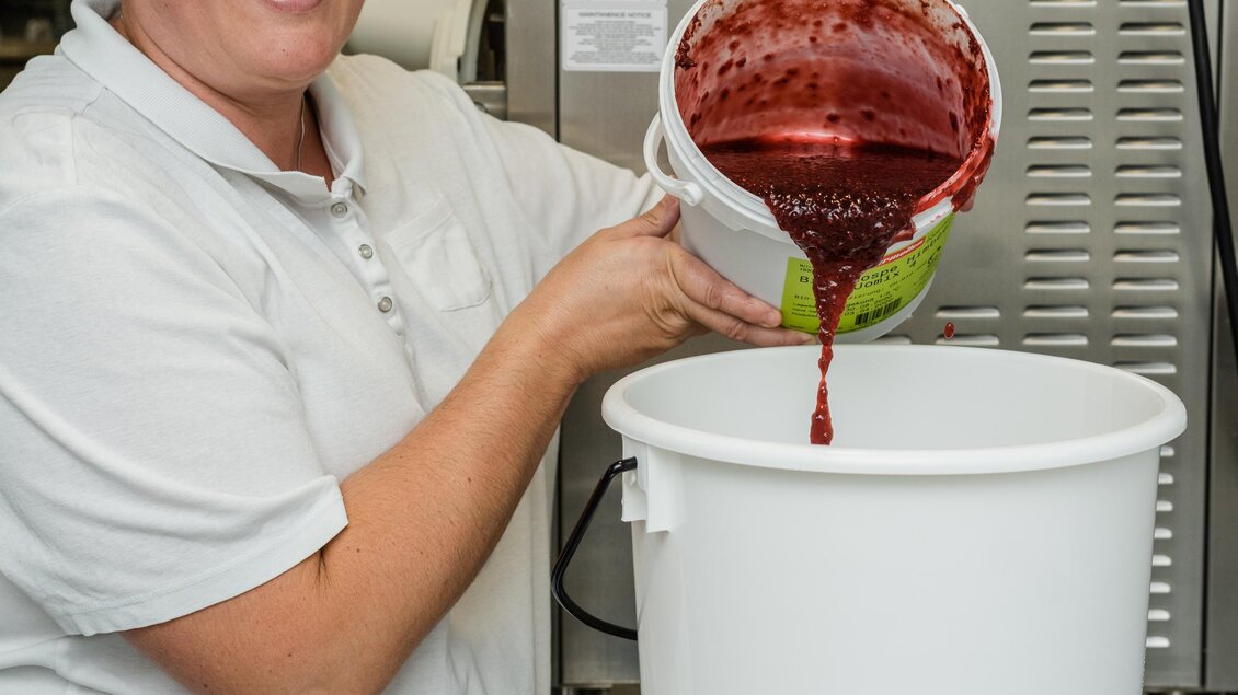 Eine Mitarbeiterin gießt eine rote Flüssigkeit in einen großen Behälter. Sie trägt eine Haube und ein einheitliches Shirt in einer modernen Küche. | © Martina Siebenhandl