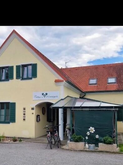 Ein gelbes Haus mit grünem Fensterladen und rotem Dach. Es gibt ein Fahrrad vor der Tür und einen kleinen Garten. | © Janisch
