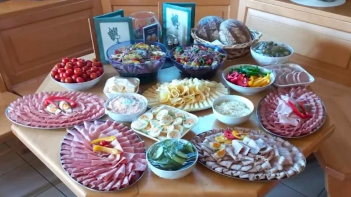 Ein großer Tisch mit verschiedenen Körben und Schalen voller frischer Lebensmittel. Es gibt Wurst, Käse, Gemüse, einen bunten Salat und Tomaten, die zusammen ein einladendes Buffet bilden. | © Janisch
