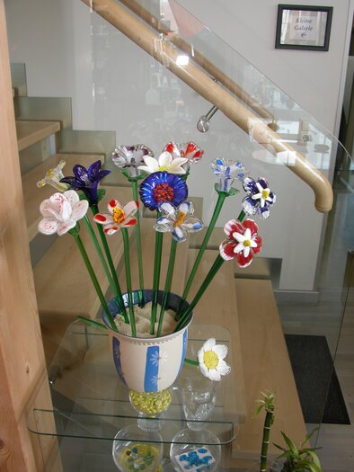 A blooming arrangement of colorful artificial flowers in a vase. The flowers are situated in a modern room with an elegant staircase railing. | © Glas Peyrer