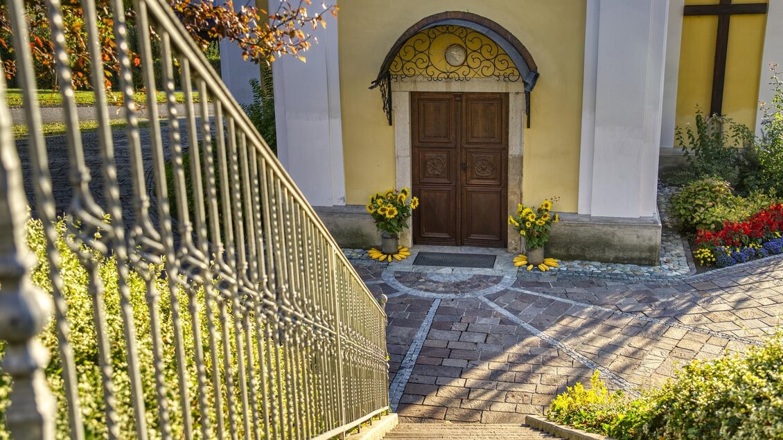 Ein schöner Eingang zu einem Gebäude mit einer Holztür und bunten Blumenkübeln. Eine gewundene Treppe führt zu dem Eingang. | © Kurkommission Bad Blumau