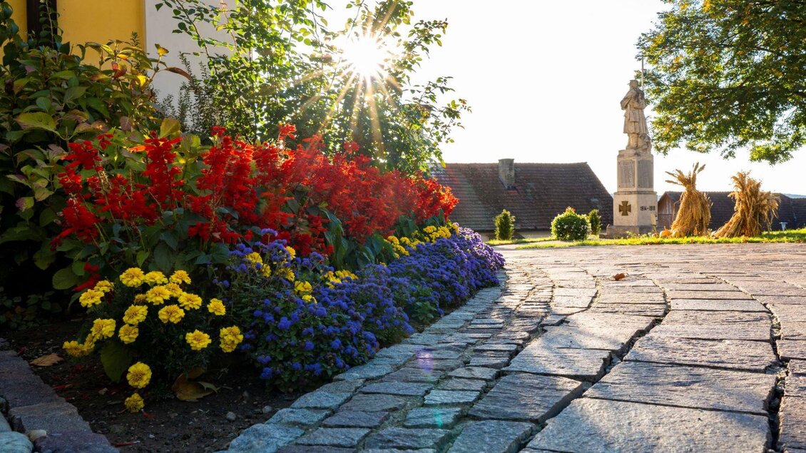 Ein schöner Gartenweg mit bunten Blumen in Rot, Gelb und Blau. Im Hintergrund steht eine Kirche unter einem klaren Himmel. | © Kurkommission Bad Blumau