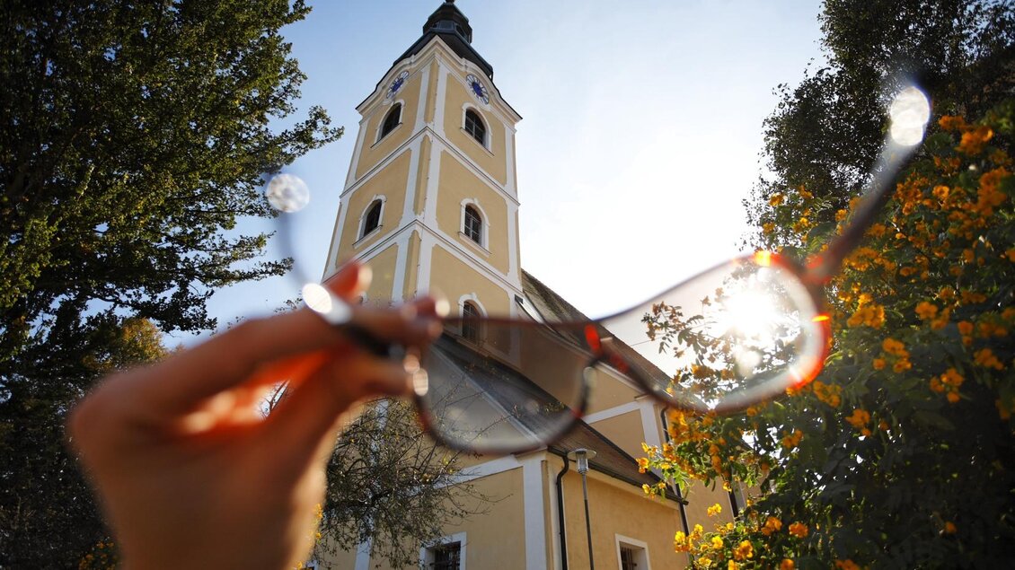 Ein Hand zeigt eine Sonnenbrille vor einer Kirche mit einem hohen Turm. Die Sonne scheint hell im Hintergrund und der Himmel ist klar. | © Sonja Hofinger