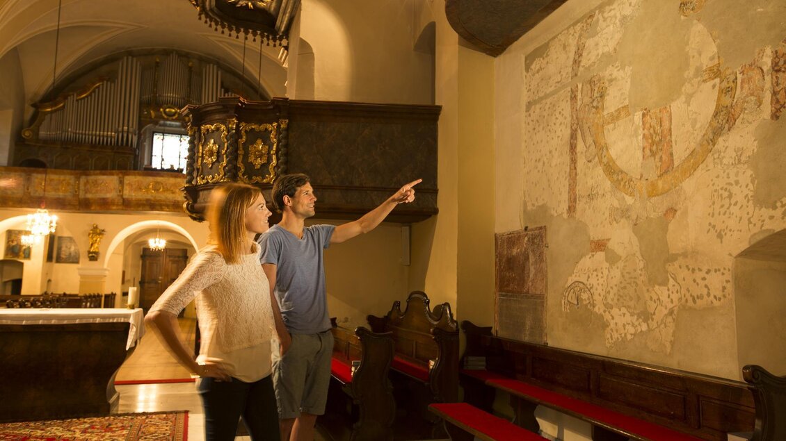 Ein Paar steht in einer Kirche und betrachtet eine Wandmalerei. Sie zeigen auf die Kunstwerke, während die Atmosphäre gedämpft und einladend ist. | © Sonja Hofinger
