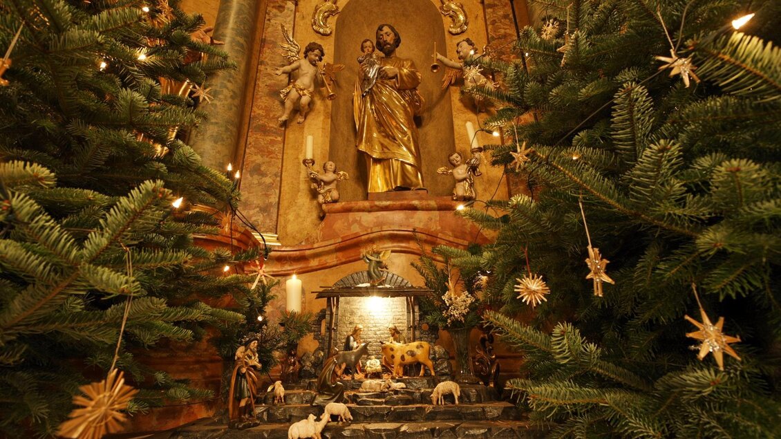 Eine festliche Krippe mit Figuren in einer Kirche, umgeben von Weihnachtsbäumen. Im Hintergrund steht eine goldene Statue. | © Sonja Hofinger