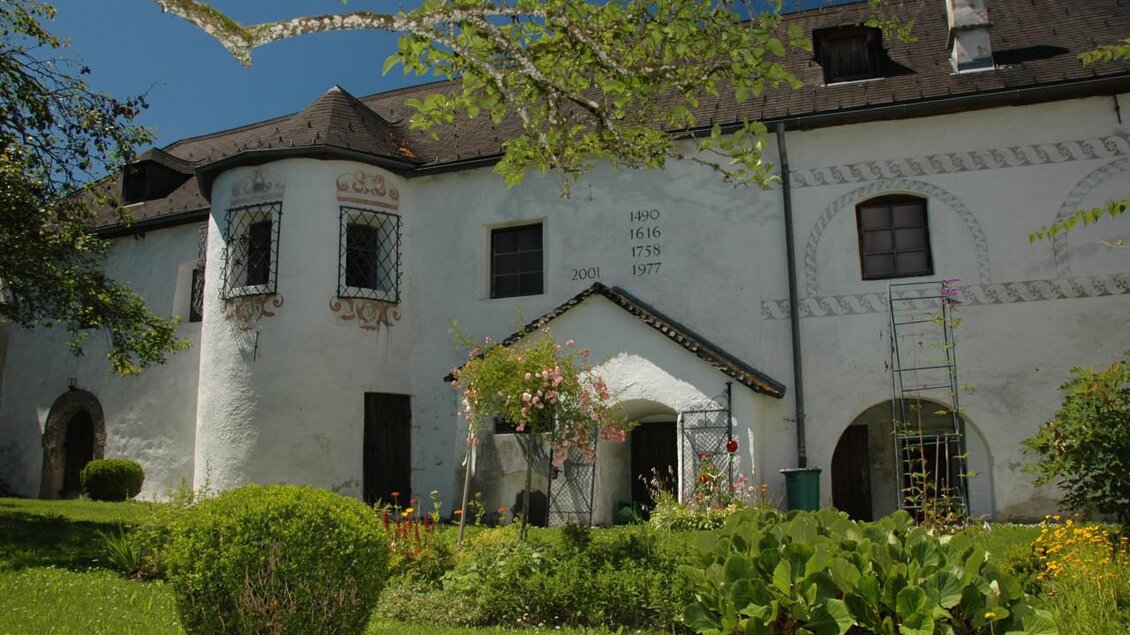 Ein historisches Gebäude mit weißer Fassade und geschmackvollen Fenstern. Der Garten ist grün und blühend, umgeben von verschiedenen Pflanzen. | © TVB Haus-Aich-Gössenberg