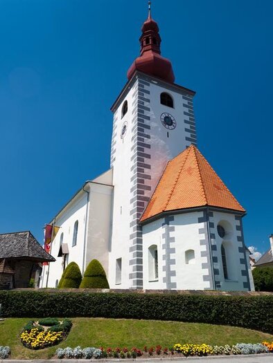 Eine malerische Kirche mit einem roten Turm und einem spitzen Dach. Umgeben von gepflegten Gärten und bunten Blumen. | © Harry Schiffer