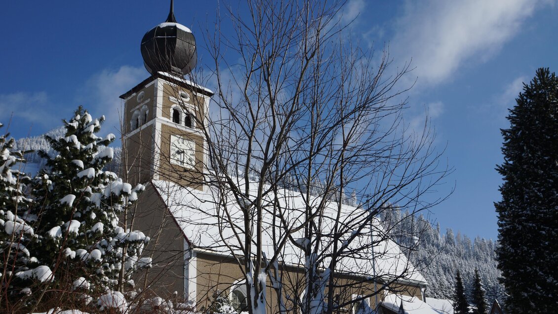 Eine malerische Kirche im Schnee mit einem hohen Kirchturm. Der Himmel ist klar und blau, umgeben von schneebedeckten Bäumen. | © Alois Leitner