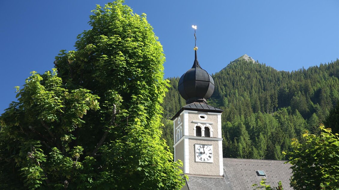 Ein Kirchturm mit einer charakteristischen Zwiebelhaube und einer Uhr ist von grünen Bäumen umgeben. Im Hintergrund sind sanfte Hügel und ein blauer Himmel zu sehen. | © Erlebnisregion Murtal