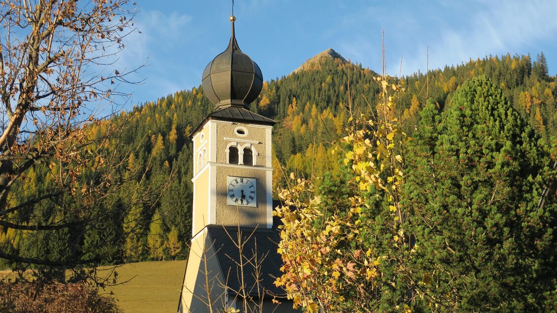 Ein Kirchturm mit einer charakteristischen Kuppel steht vor einer bewaldeten Berglandschaft. Die Bäume zeigen herbstliche Farben im Hintergrund. | © Marlene Zandl