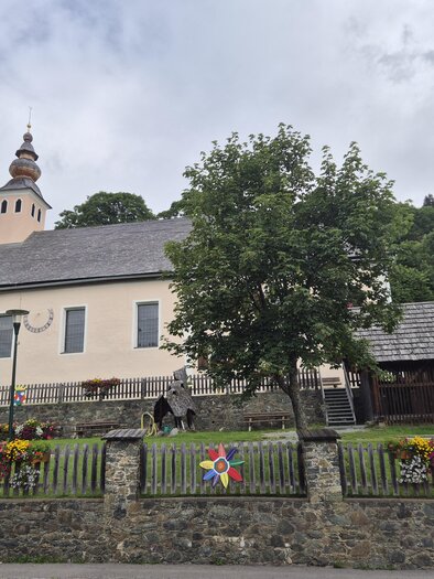 Eine hübsche Kirche mit einem Turm und einem geschützten Vorplatz. Im Vordergrund befindet sich eine Steinmauer mit bunten Blumen. | © Tourismusverband Murau