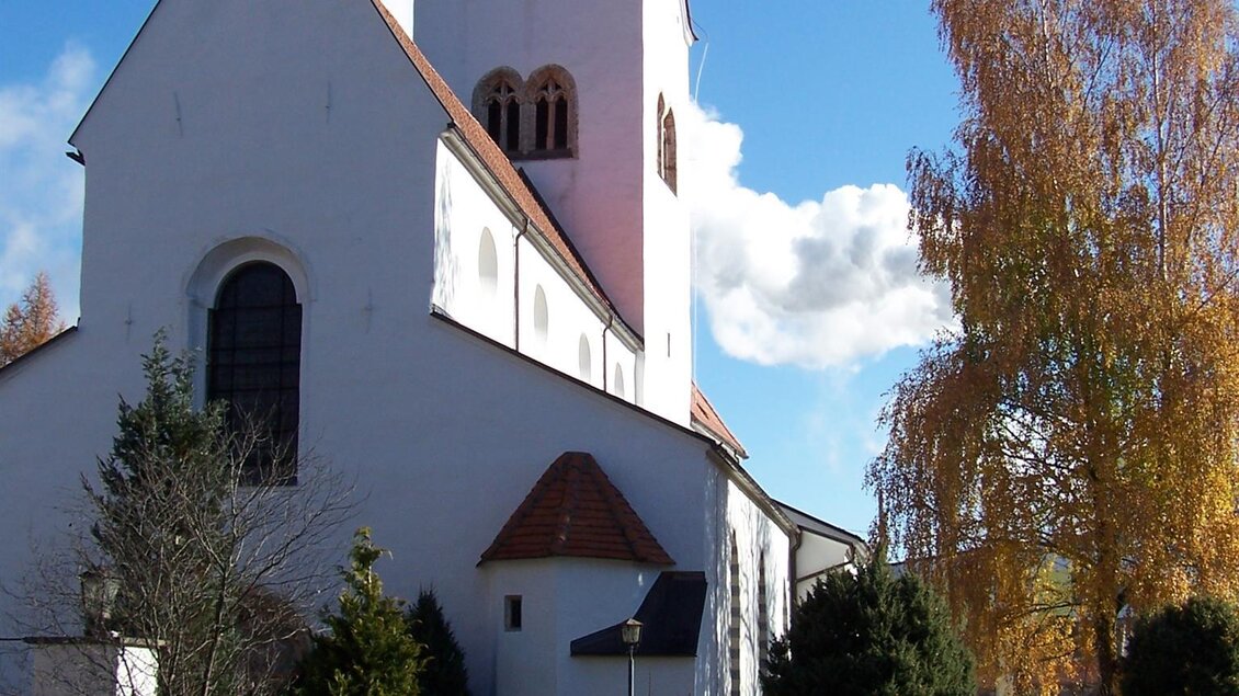 Eine weiße Kirche mit einem roten Dach und einem hohen Turm. Im Hintergrund stehen bunte Bäume und der Himmel ist klar und blau. | © Kth. Kirche Hl. Nikolaus Pöls