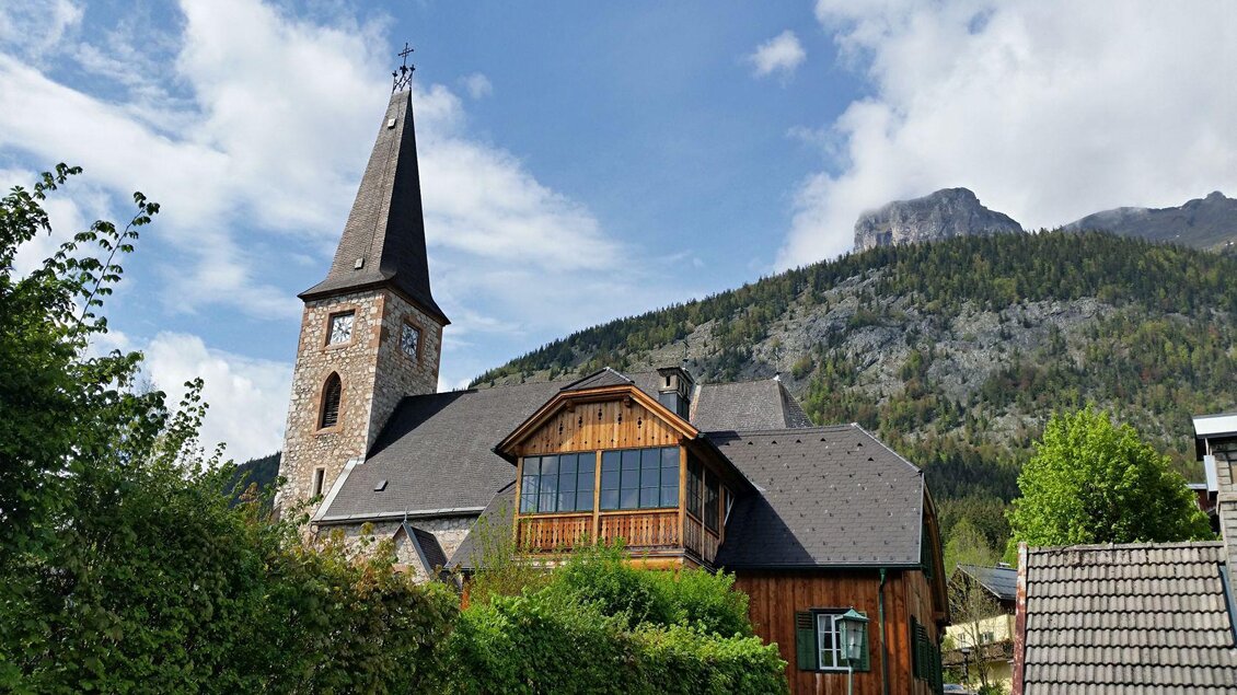 Ein malerisches Dorf mit einer Kirche und traditionellen Holzgebäuden. Im Hintergrund sind majestätische Berge und ein blauer Himmel zu sehen. | © Rabensteiner