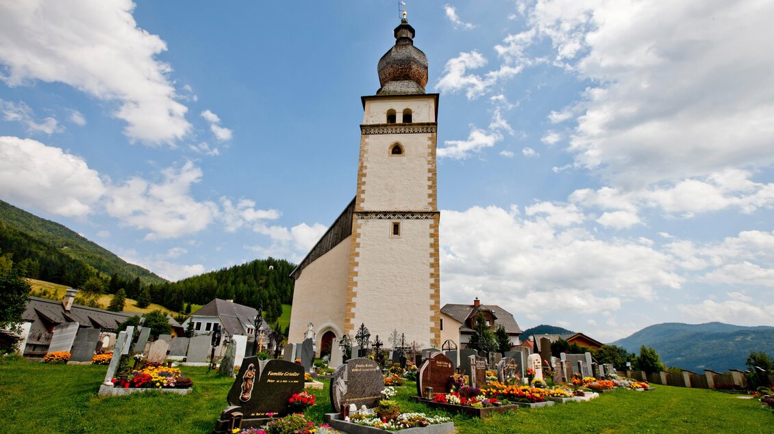 Eine Kirche mit einem hohen Turm steht inmitten eines Friedhofs. Im Hintergrund sind grüne Hügel und ein blauer Himmel mit Wolken zu sehen. | © Holzwelt Murau