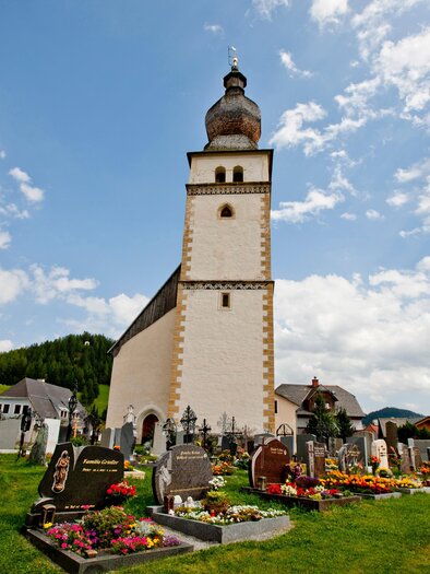 A church with a tall steeple stands in the middle of a cemetery. In the background, there are green hills and a blue sky with clouds. | © Holzwelt Murau