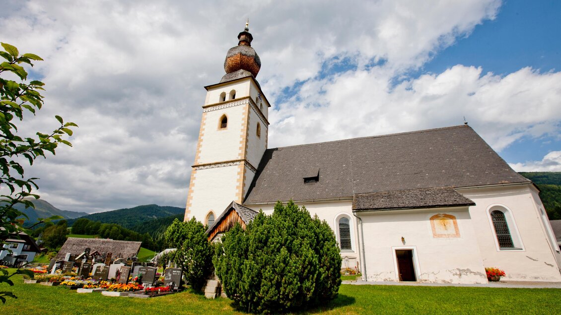Eine schöne Kirche mit einer markanten Glockenkuppe und umgeben von einer grünen Wiese. Im Vordergrund ist ein Friedhof mit bunten Blumen zu sehen. | © Holzwelt Murau
