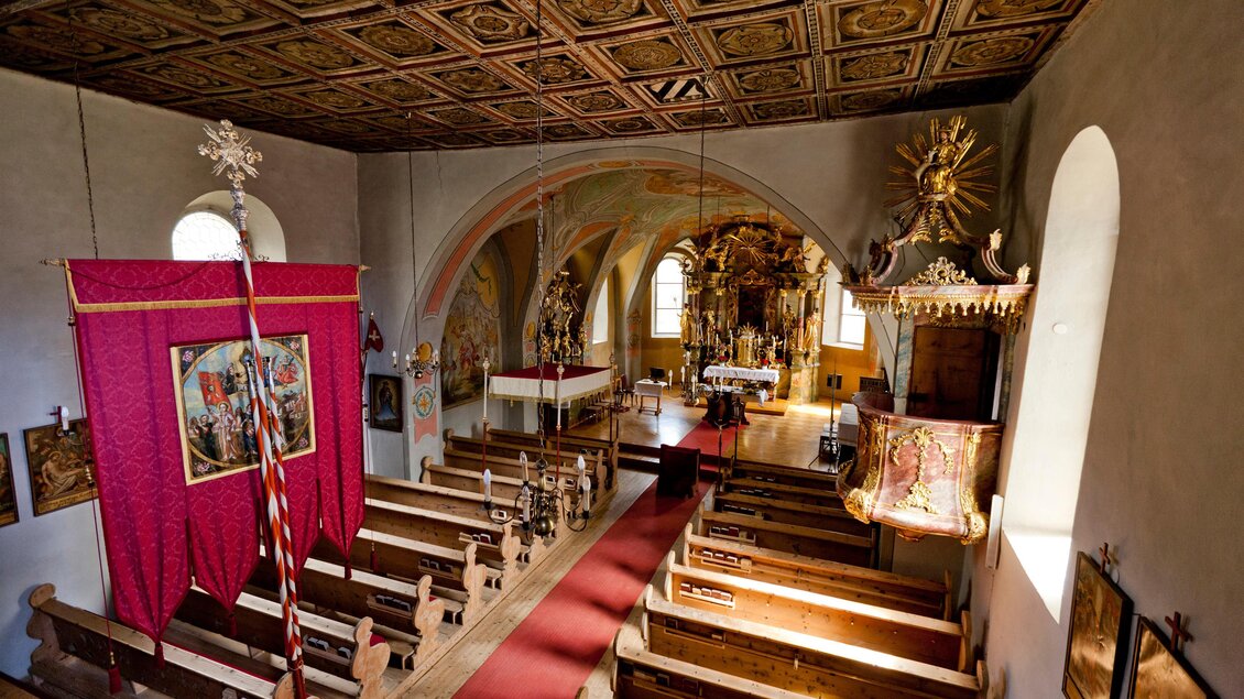 Eine Innenansicht einer Kirche mit Holzbänken und einem bunten Altar. Der Raum hat eine schöne Decke und ist mit religiösen Kunstwerken dekoriert. | © Holzwelt Murau