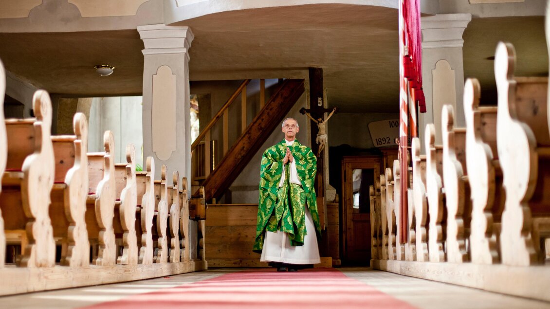 Ein Priester steht in einer Kirche auf einem roten Teppich. Die Bänke sind aus Holz und ein Kreuz ist im Hintergrund sichtbar. | © Holzwelt Murau