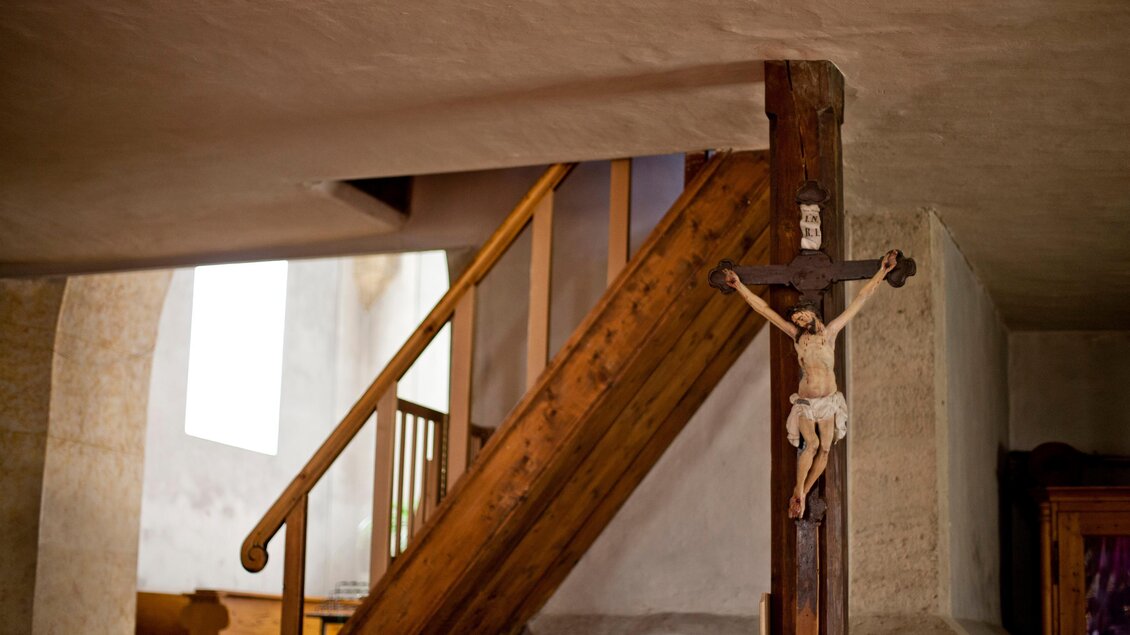 Ein gemütlicher Innenraum mit einer Holztreppe und einem Kreuz an der Wand. Das Licht fällt sanft durch ein Fenster und schafft eine ruhige Atmosphäre. | © Holzwelt Murau