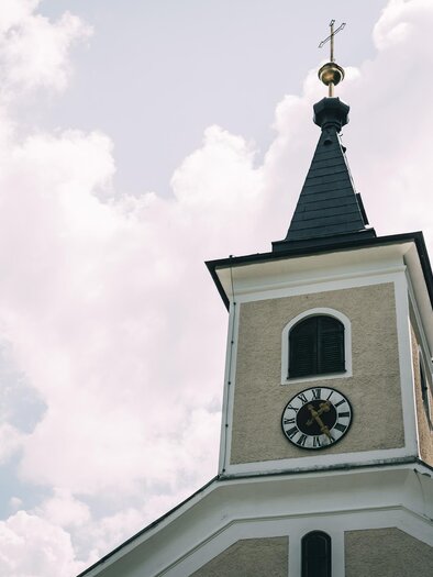 A church tower with a clock and a cross at the top. The sky is bright and cloudy. | © Armin Walcher