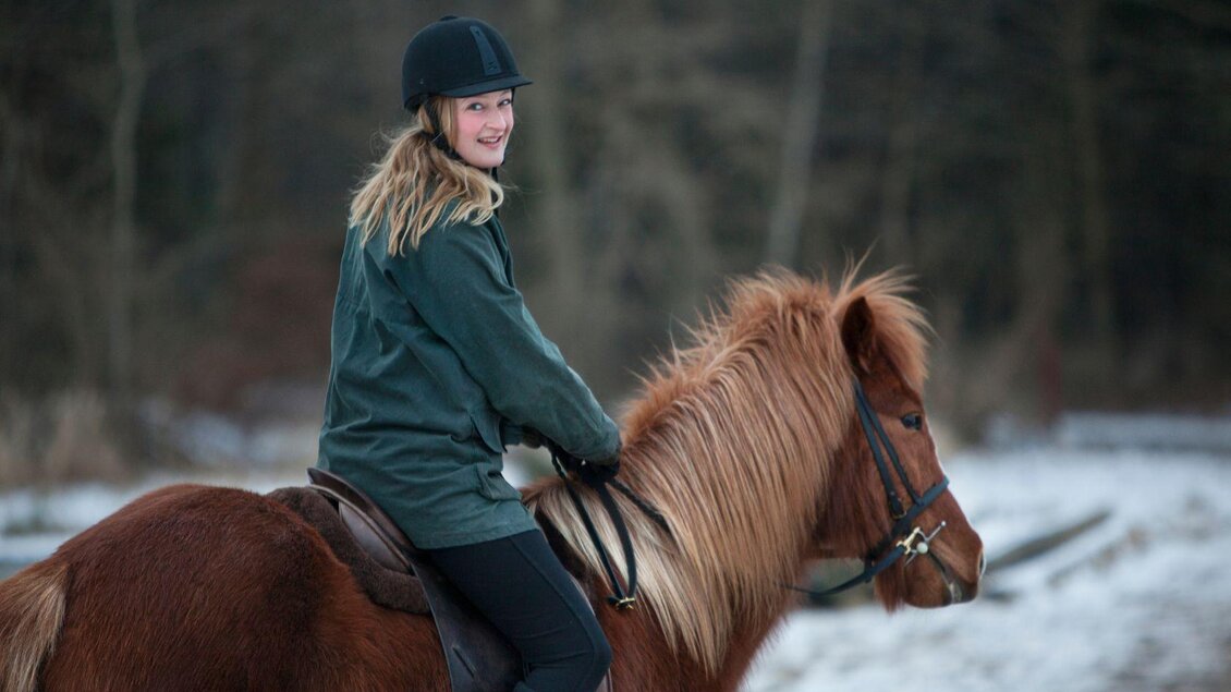 Ein Mädchen sitzt auf einem braunen Pferd in einer winterlichen Landschaft. Sie lächelt und trägt eine Reitkappe sowie eine warme Jacke. | © Pferdefreunde Zloam - www.pferdefreunde-zloam.at