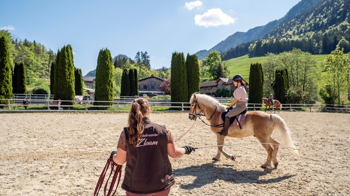 Eine Reitstunde auf einem Platz im Freien. Eine Person führt ein Pferd, während eine andere Person auf dem Pferd sitzt und reitet. | © www.zloam.at