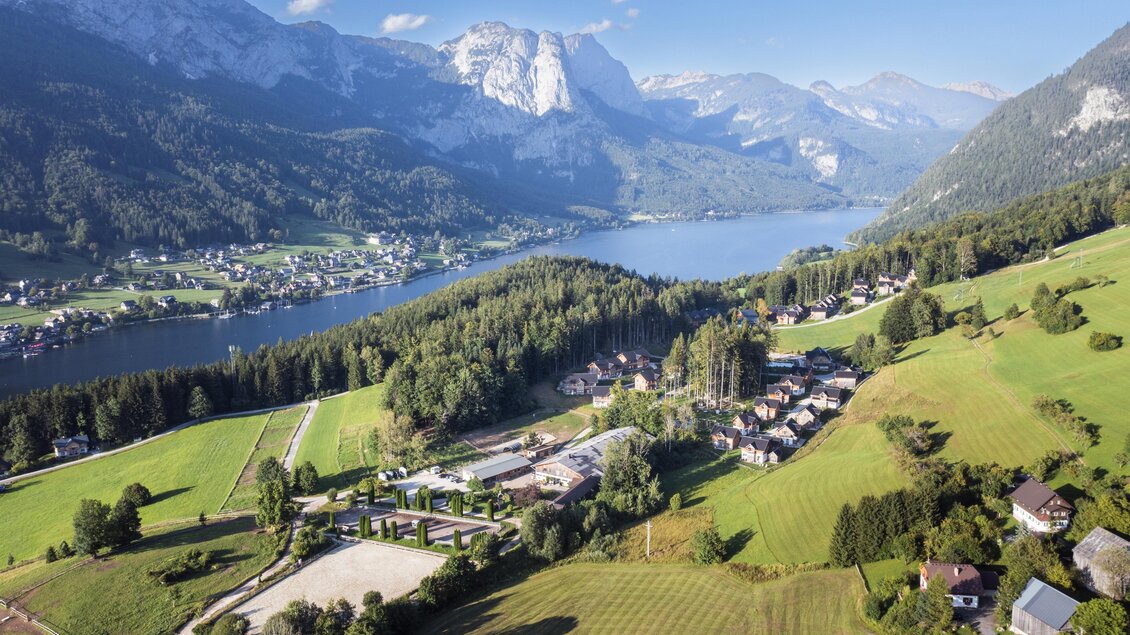 Eine malerische Landschaft mit Bergen, einem klaren See und üppigen grünen Wiesen. Im Vordergrund sind kleine Häuser und landwirtschaftliche Flächen zu sehen. | © www.zloam.at