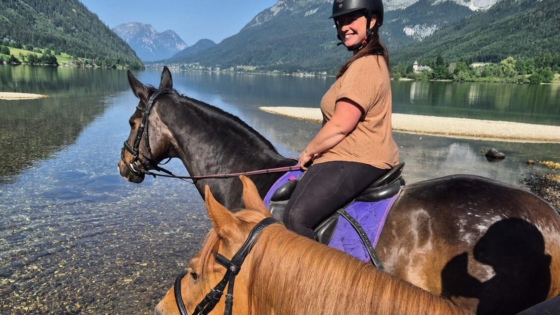 Eine Frau sitzt auf einem schwarzen Pferd am Ufer eines klaren Sees. Im Hintergrund sind Berge und eine grüne Landschaft zu sehen. | © www.zloam.at