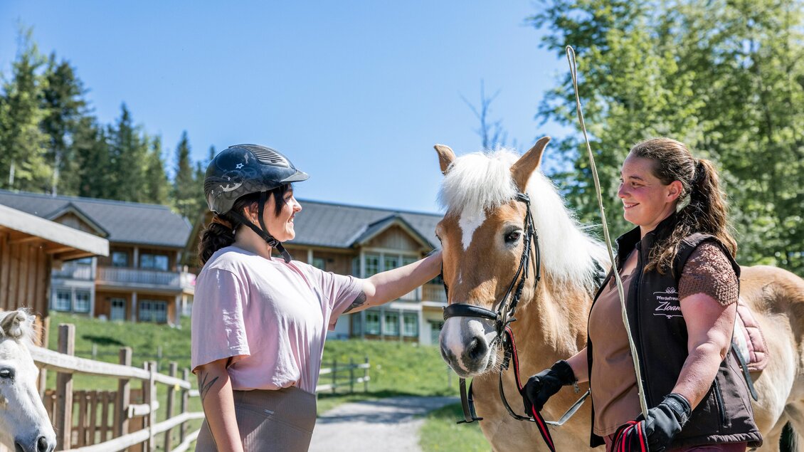 Zwei Frauen stehen im Freien und interagieren mit einem Pferd. Im Hintergrund sind Gebäude und Bäume zu sehen. | © www.zloam.at